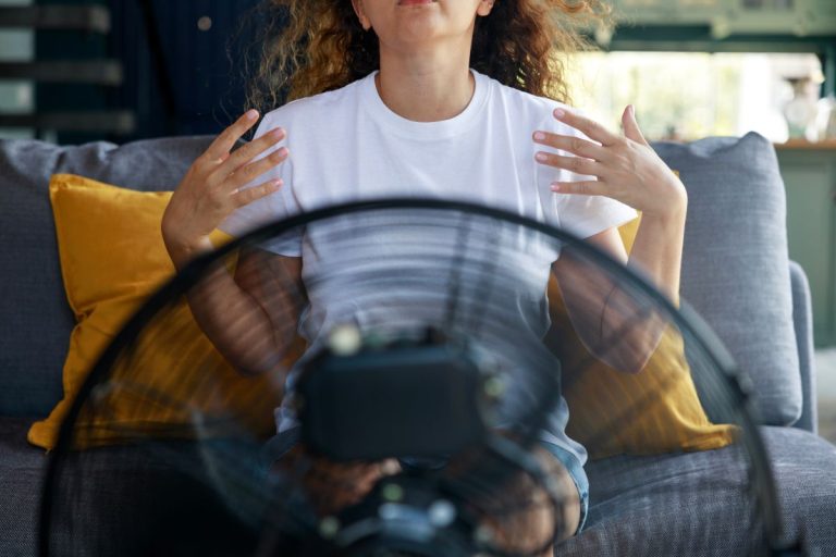Woman trying to cool herself with electric fan during heatwave at home